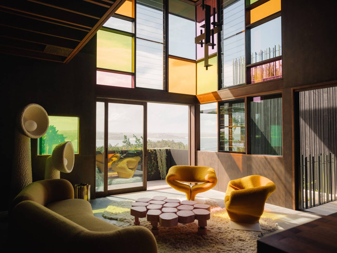 Gio sofa in the double-height living room of Holocene House, with sculptural seating and colourful glass windows.