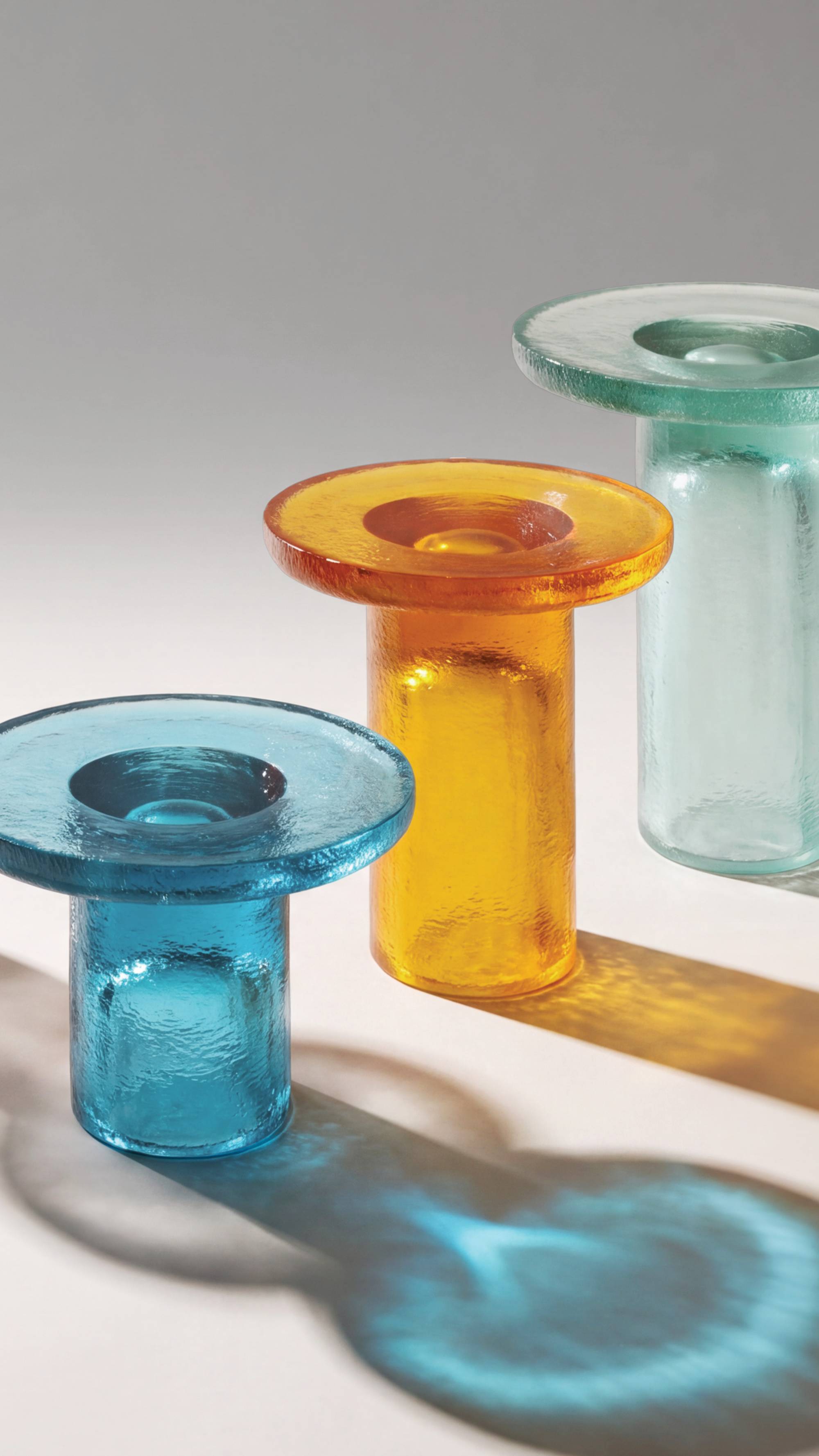 Five Momo resin side tables in rust, clear, light blue, saffron and Aqua green finishes, arranged on a light background with strong sunlight and colorful translucent shadows.
