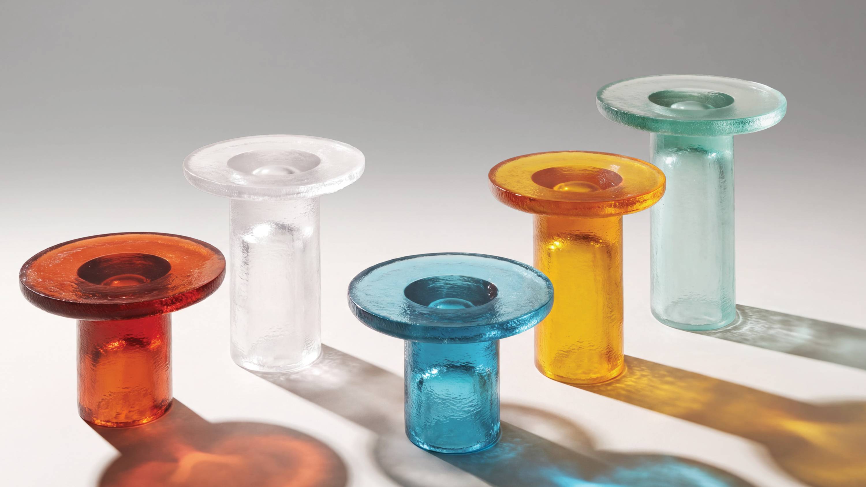 Five Momo resin side tables in rust, clear, light blue, saffron and Aqua green finishes, arranged on a light background with strong sunlight and colorful translucent shadows.